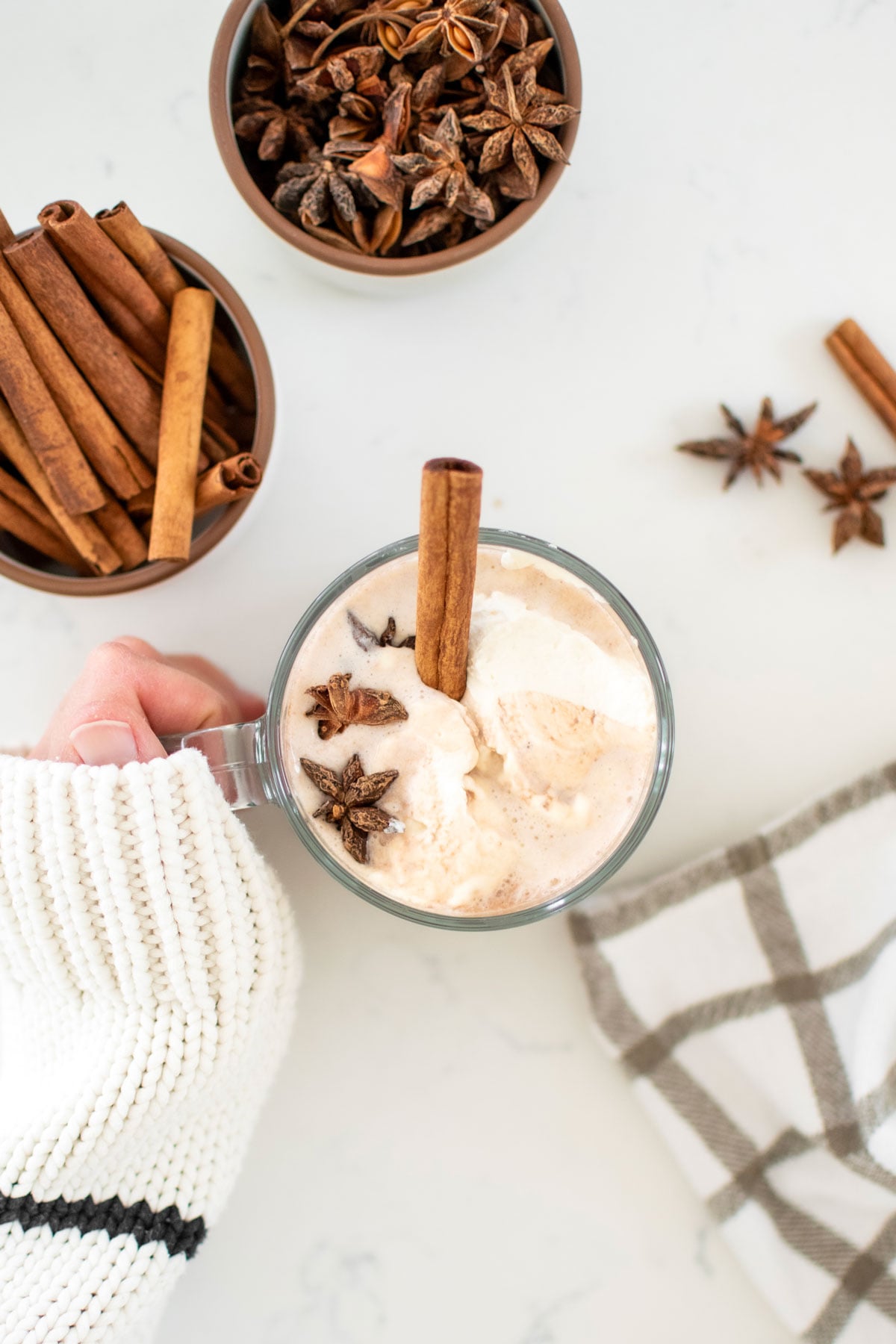 woman holding a chai tea hot chocolate in a glass mug with whipped cream, cinnamon stick and star anise.