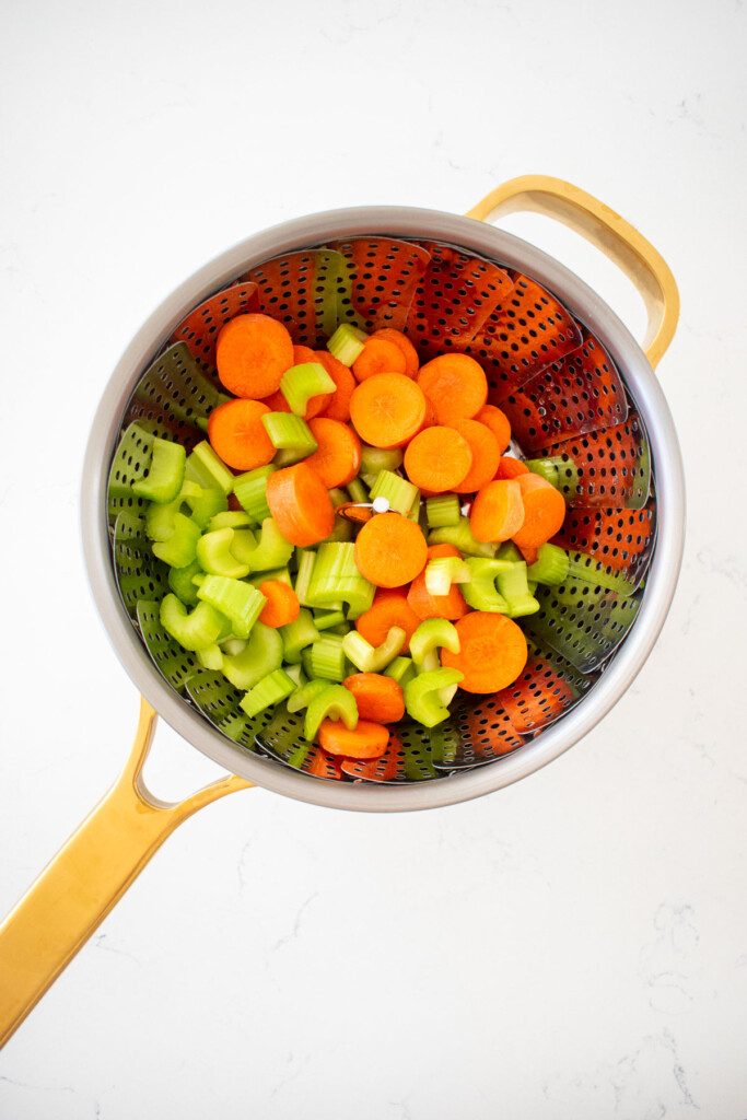 cut carrots and celery in a silver pot inside a steaming basket on a white marble counter.