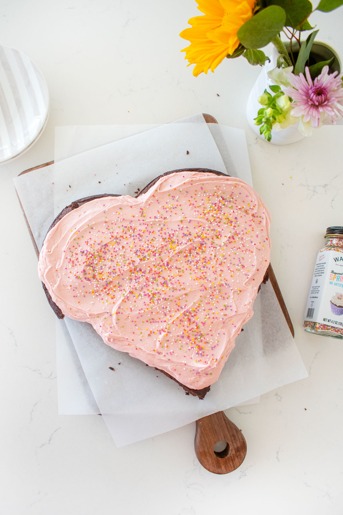pink heart shaped cake with rainbow sprinkles on a white marble counter with fresh flowers.