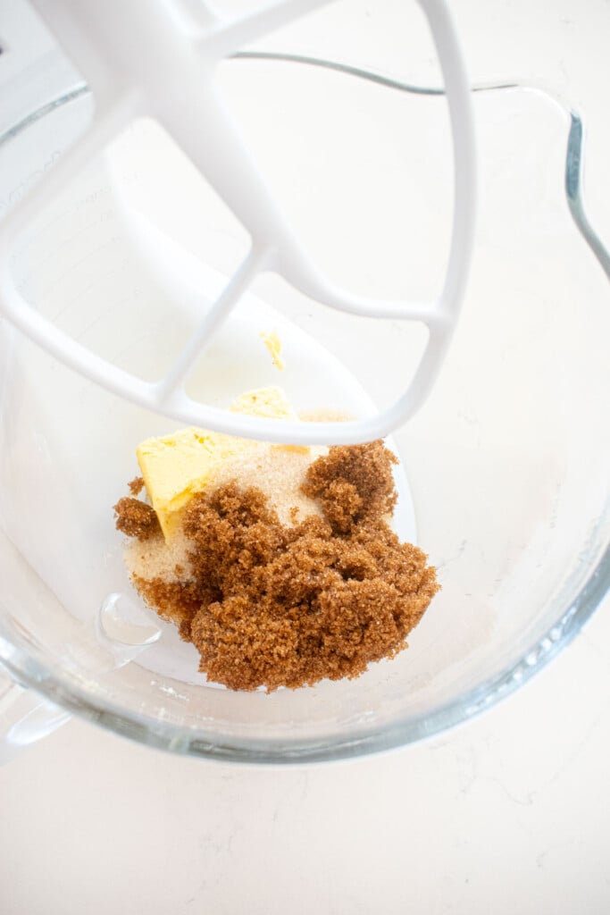 butter, cane sugar and brown sugar in a glass stand mixing bowl on a white marble counter.