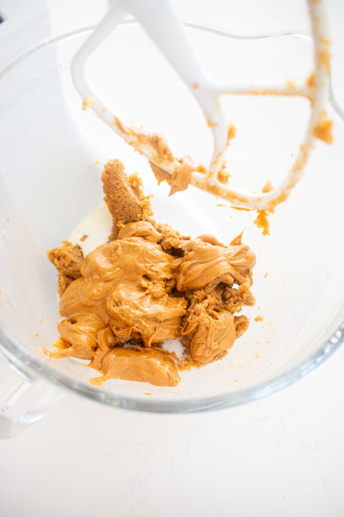 peanut butter and milk added to creamed butter and sugar in a glass stand mixing bowl on a white marble counter.