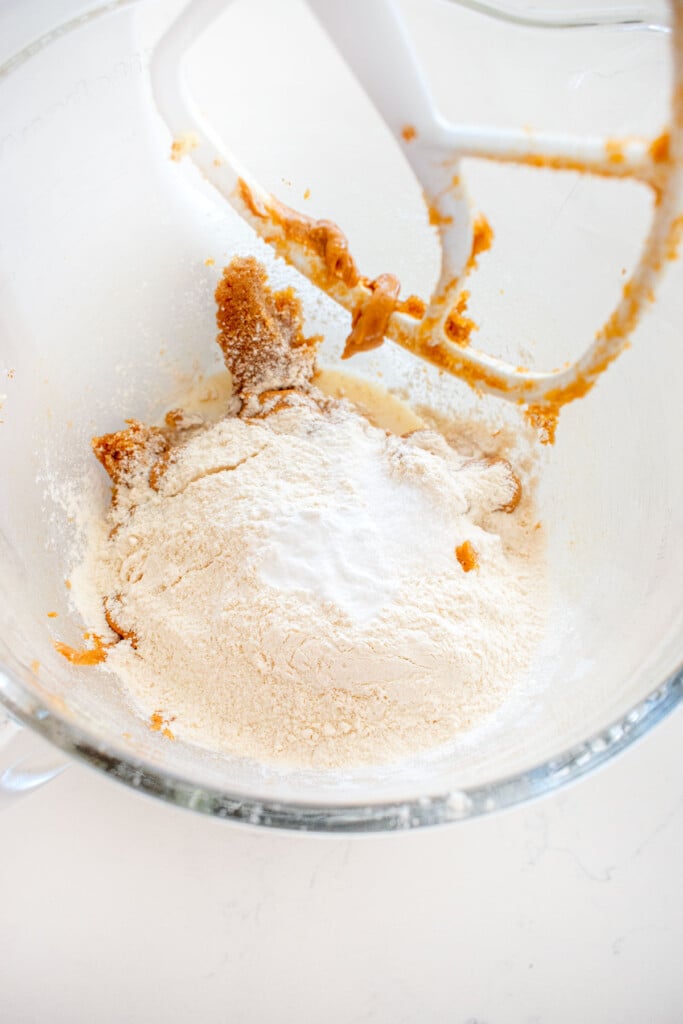 flour being added into peanut butter cookie dough in a glass stand mixing bowl on a marble counter.
