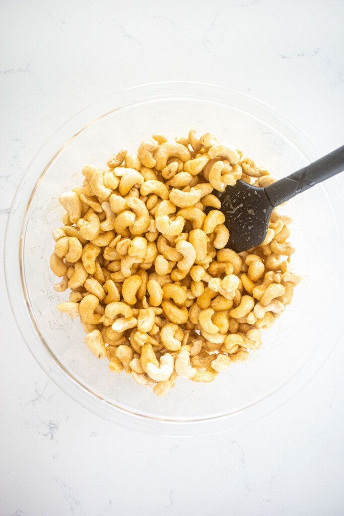cashews being stirred with honey, butter and sugar in a glass mixing bowl on a white marble countertop.