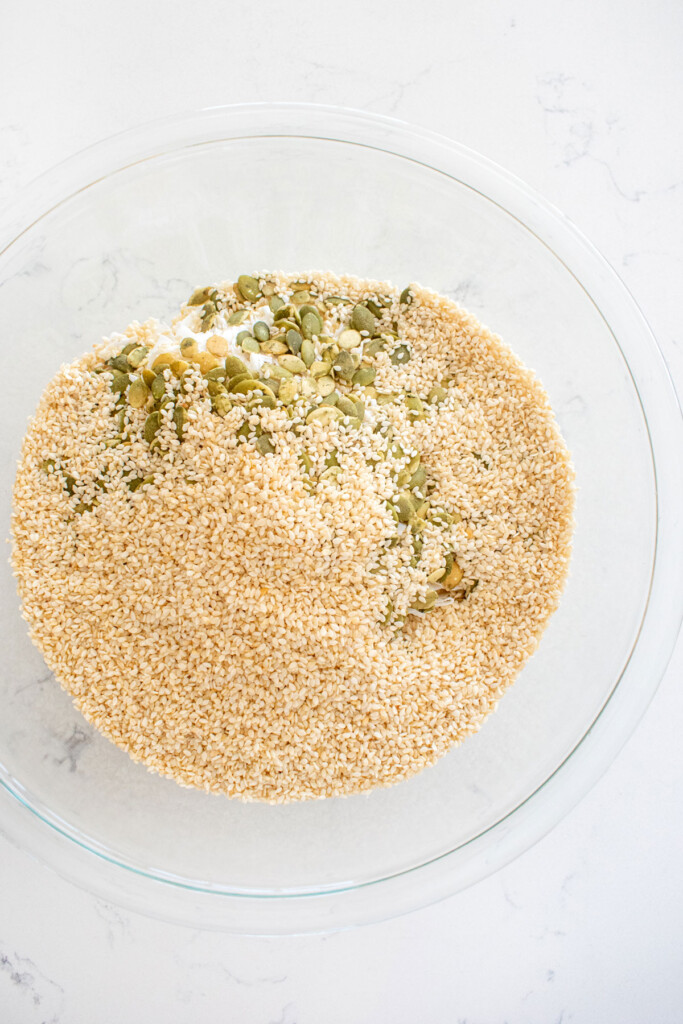 sesame seeds, pumpkin seeds and coconut in a glass mixing bowl on a white marble countertop.
