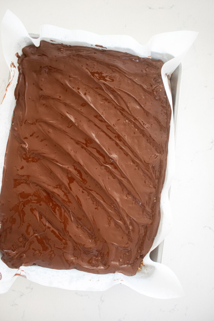 melted dark chocolate spread out onto honey sesame bark on a baking sheet on a marble countertop.