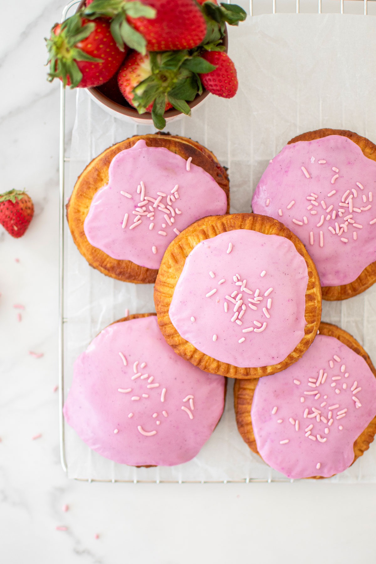 mixed berry hand pies with glaze and pink sprinkles on a cooling rack with a bowl of fresh strawberries.