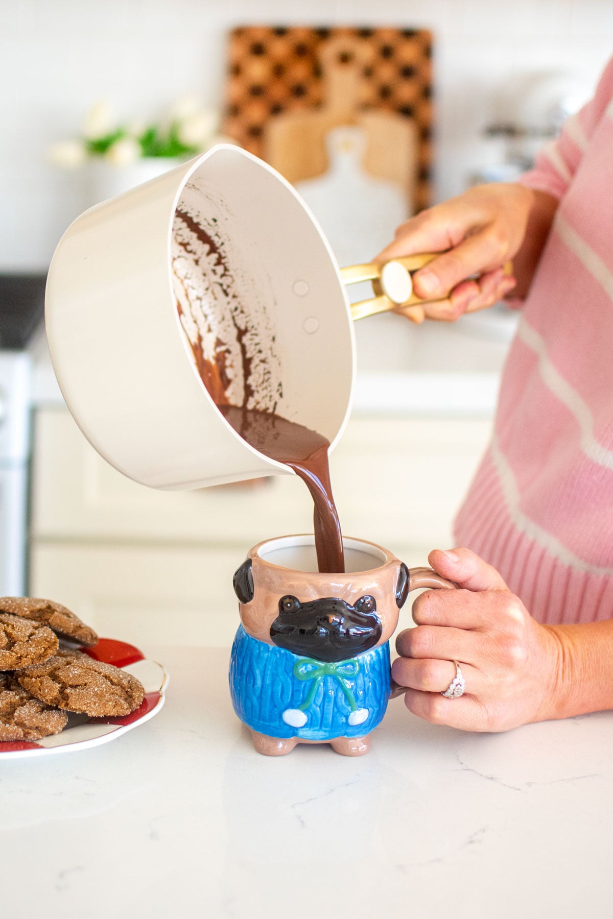 woman pouring hot chocolate from an our place pot into a pug mug at the kitchen counter.