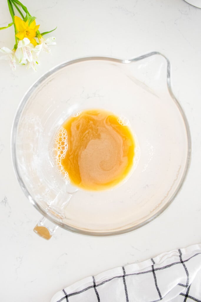 glass mixing bowl with yeast, water and maple syrup on a white marble counter.