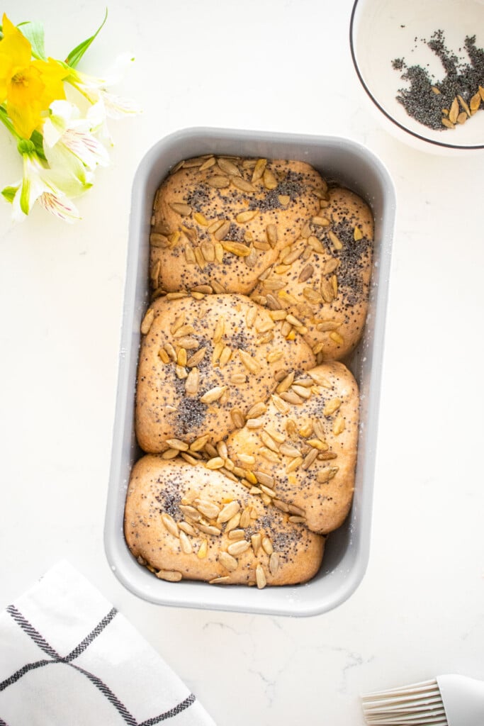 1-pound loaf pan with dough balls placed zig zag inside it with sunflower and poppy seeds on top on a white marble counter.