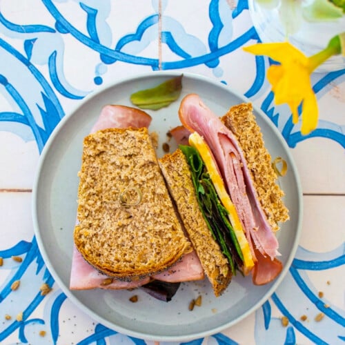 ham and cheese sandwich made with sourdough discard whole wheat sandwich bread, sliced in half on a plate on a blue tile table with fresh flowers.