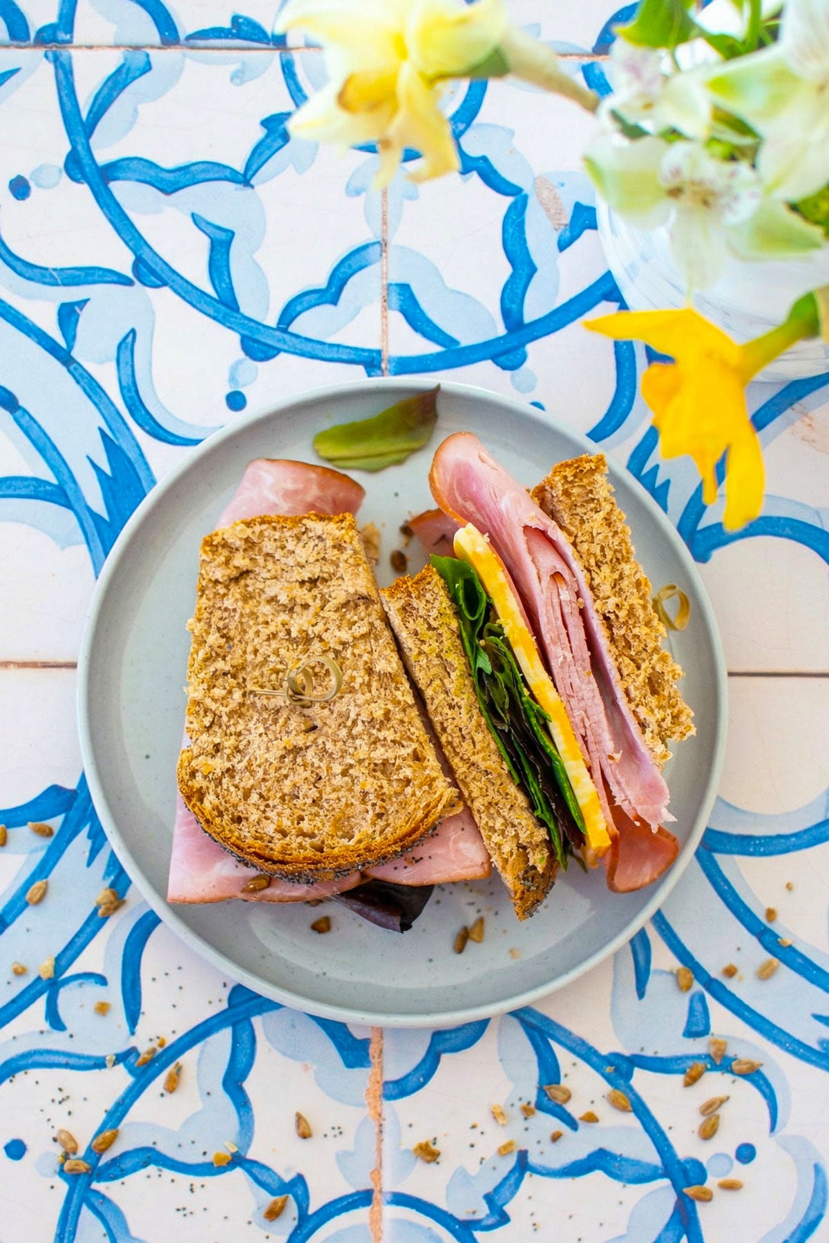 ham and cheese sandwich made with sourdough discard whole wheat sandwich bread, sliced in half on a plate on a blue tile table with fresh flowers.
