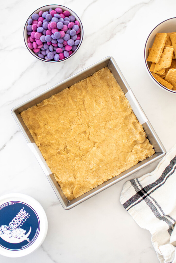 eggless cookie dough pressed evenly in a square baking pan on a white marble counter with graham crackers, pink and purple candies and marshmallow fluff.