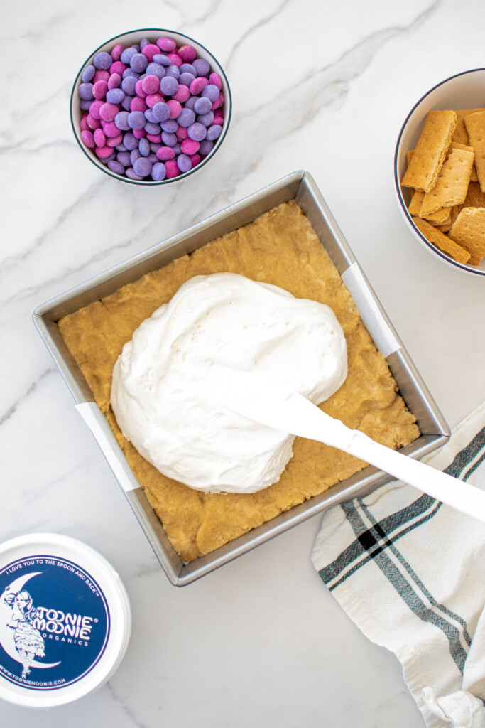 marshamllow fluff being spread onto eggless cookie dough in a square baking pan on a white marble counter.