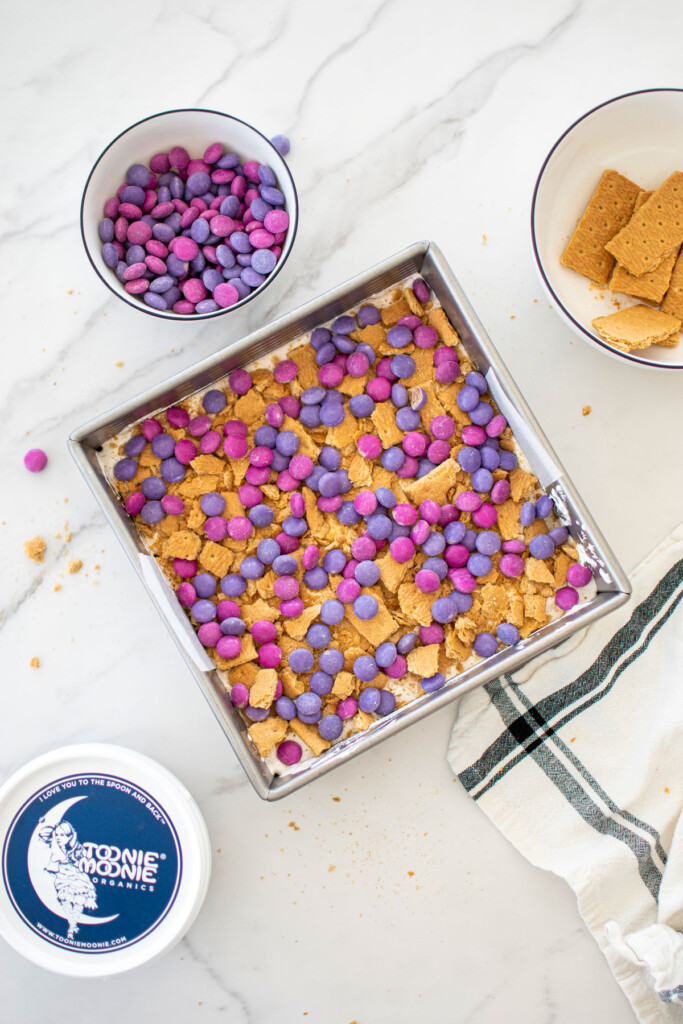 smore bars with pink and purple candies on top in a square baking pan on a white marble counter.