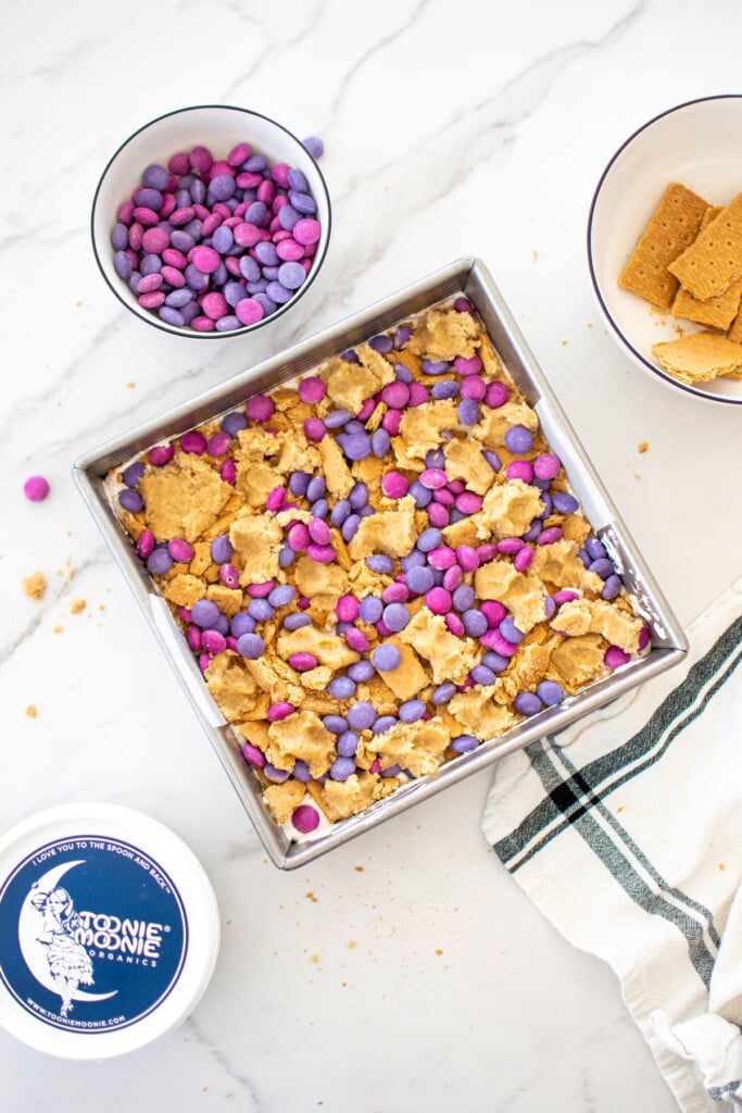 smore bars with pink and purple candies on top in a square baking pan on a white marble counter.