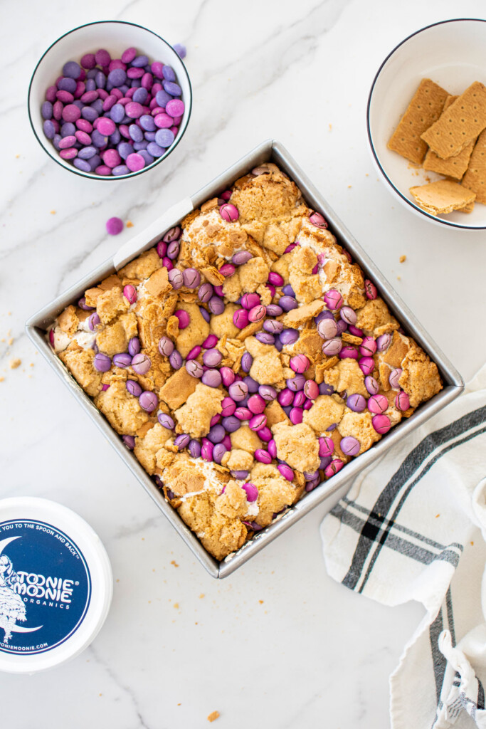 baked smore bars with pink and purple candies on top in a square baking pan on a white marble counter.