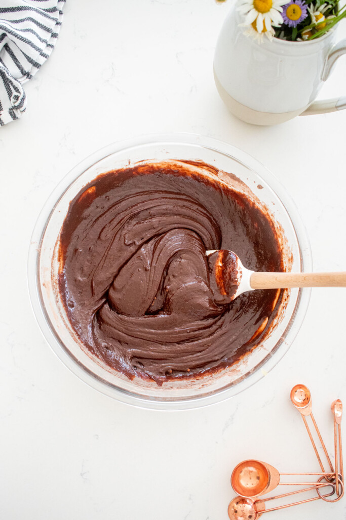 chocolate cupcake batter in a glass mixing bowl with a spatula on a marble counter with fresh flowers.