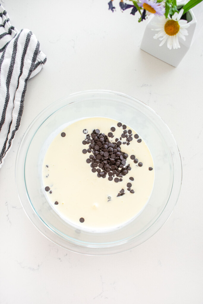glass mixing bowl with dark chocolate and heavy whipping cream on a marble counter with fresh flowers.
