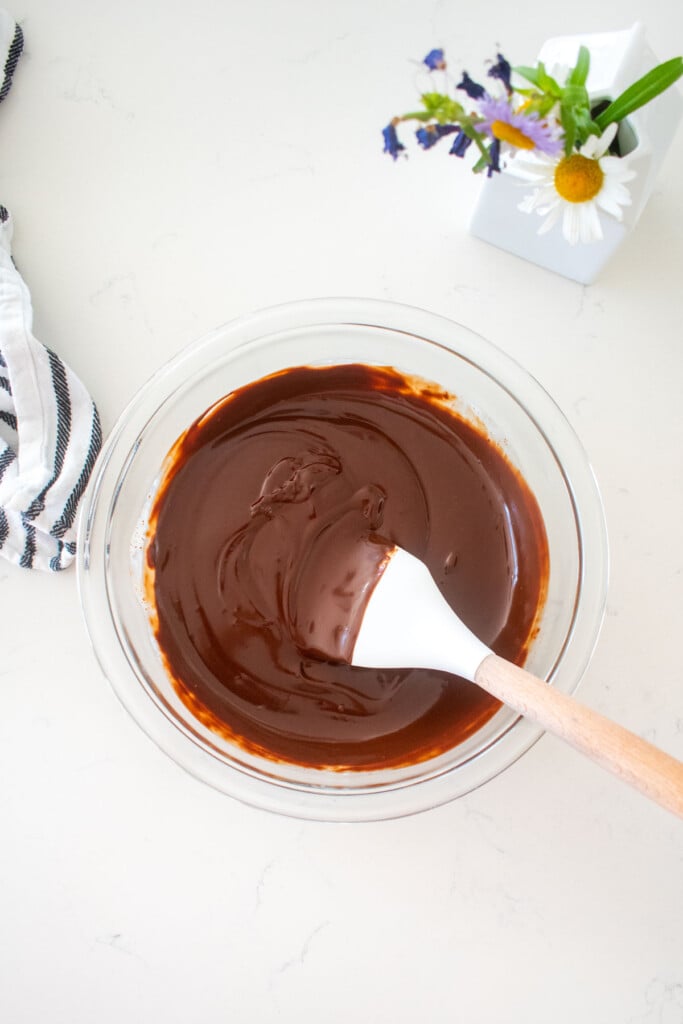 smooth chocolate ganache in a glass mixing bowl on a marble counter with a spatula in it.