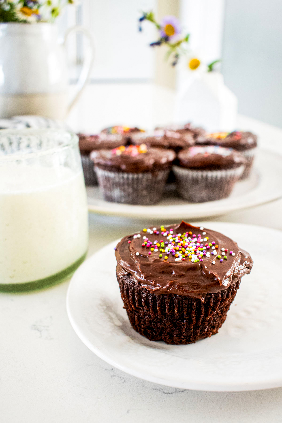 chocolate cupcake with chocolate frosting and rainbow sprinkles on a plate with a glass of milk on a kitchen counter.