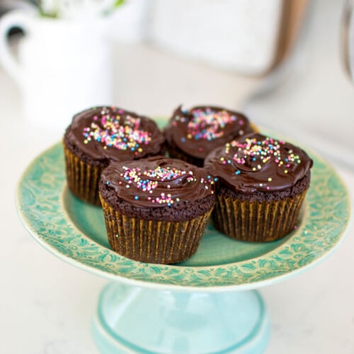 chocolate cupcakes with rainbow sprinkles on a green cake plate on a white marble counter.
