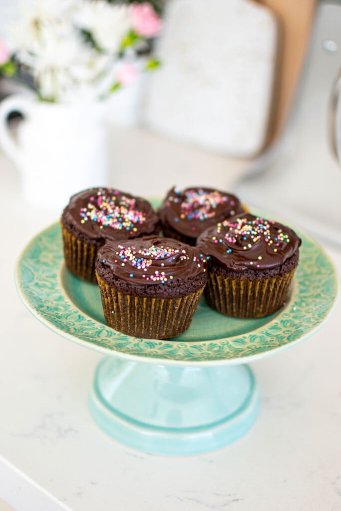 chocolate cupcakes with rainbow sprinkles on a green cake plate on a white marble counter.