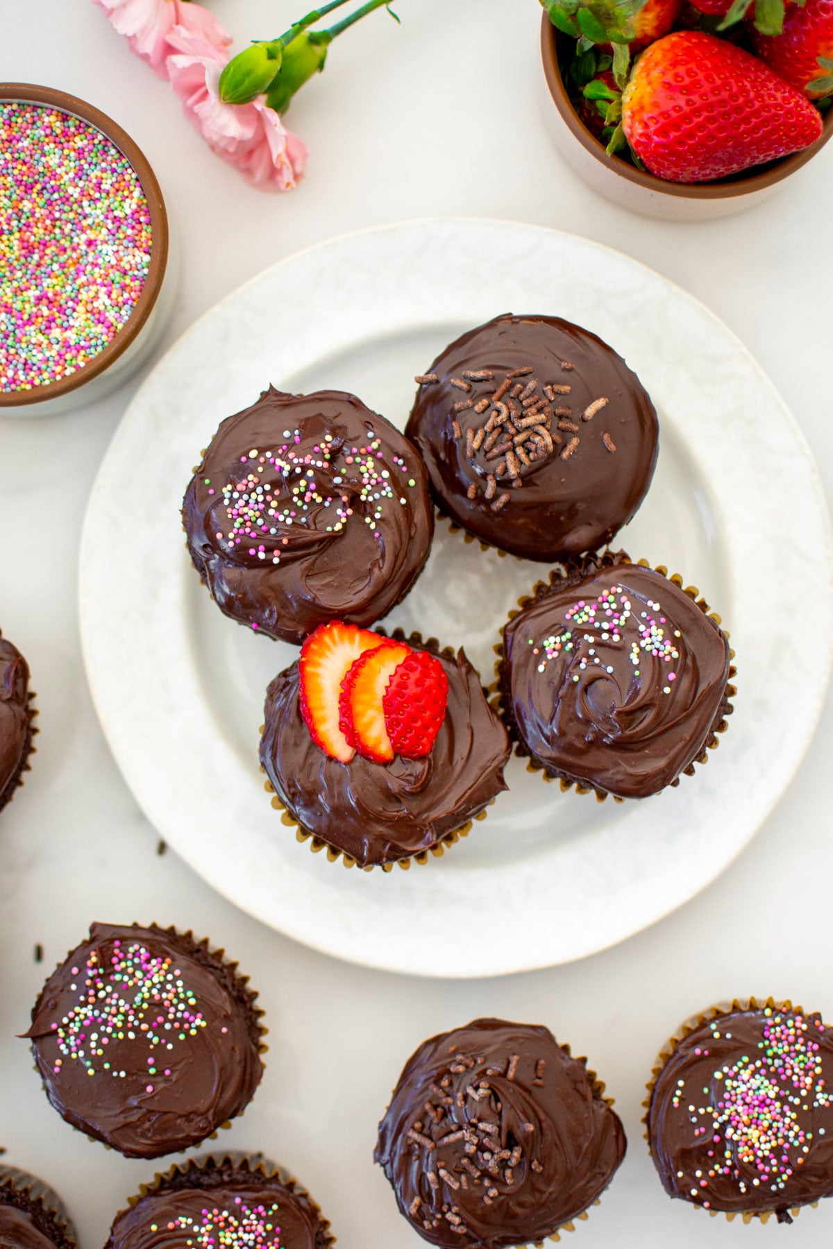 plate of chocolate cupcakes with chocolate ganache frosting and different toppings on a plate on a white marble counter.