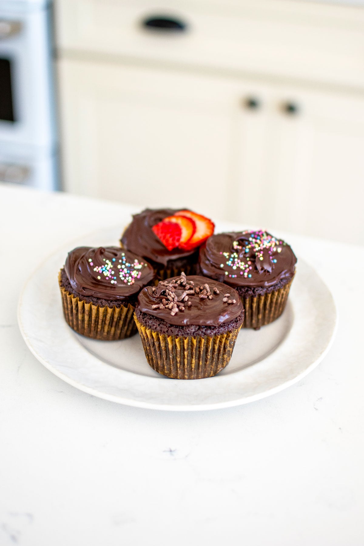 plate of chocolate cupcakes with chocolate ganache frosting with different toppings like sprinkles and strawberries on a white kitchen counter.