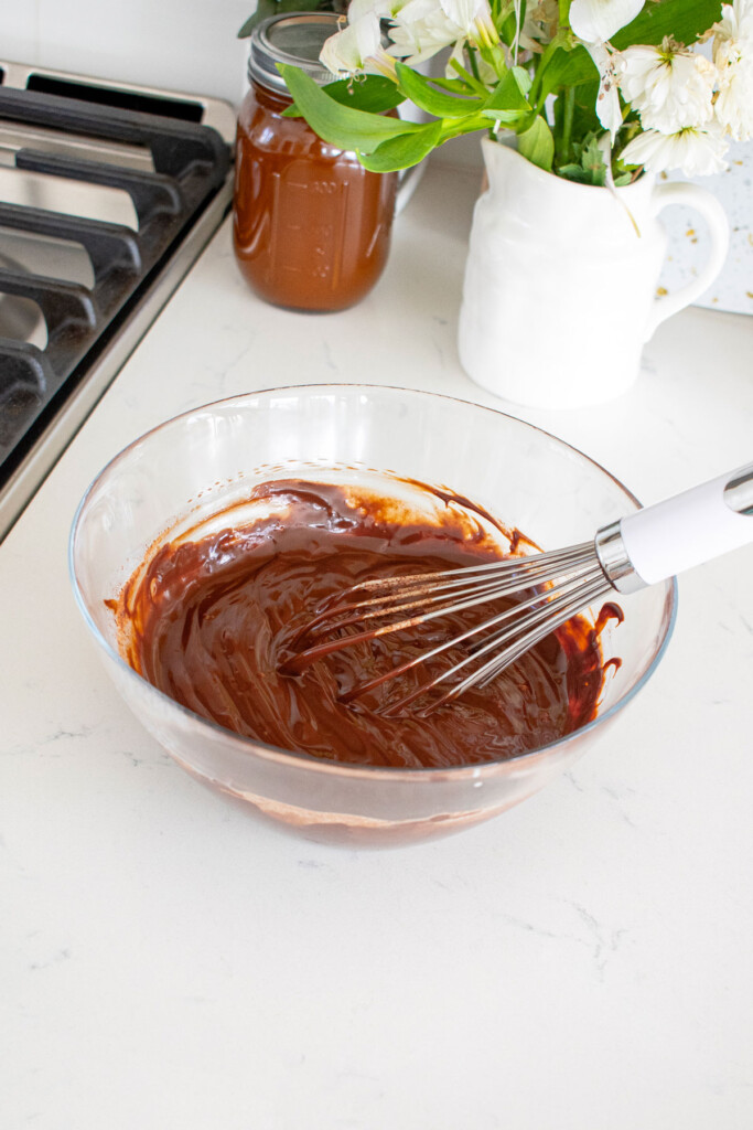 chocolate ganache in a glass mixing bowl with a whisk on a white marble counter.