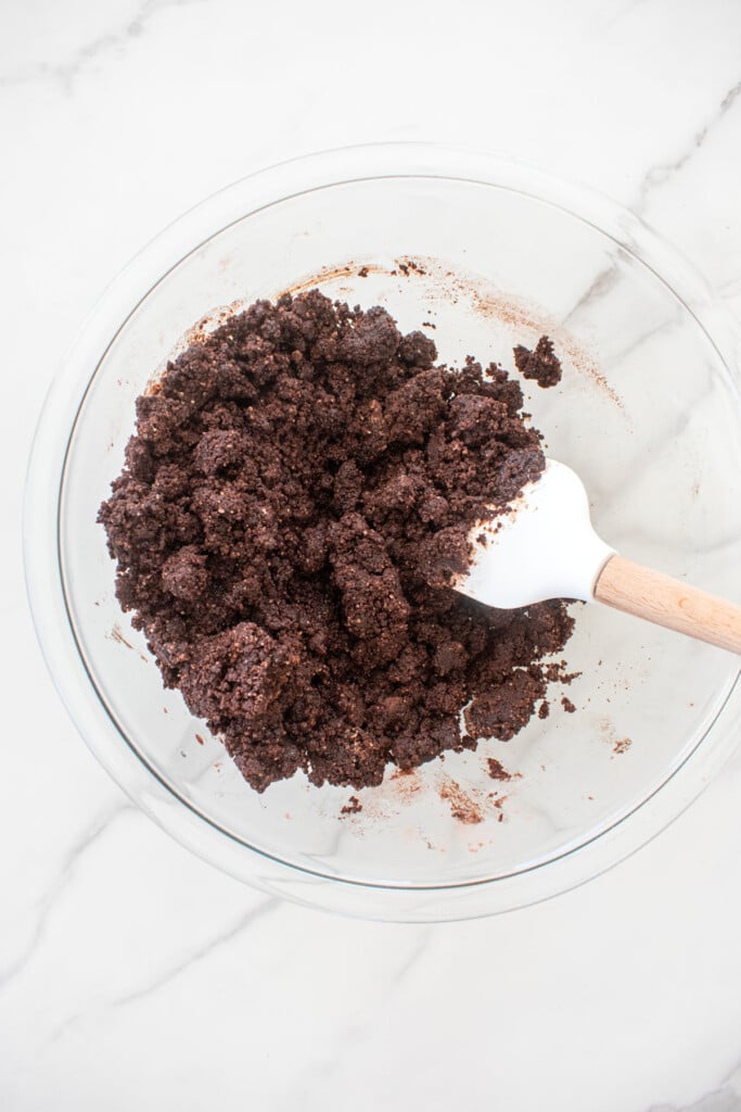crumbly chocolate shortbread dough in a glass mixing bowl with a spatula on a white marble counter.