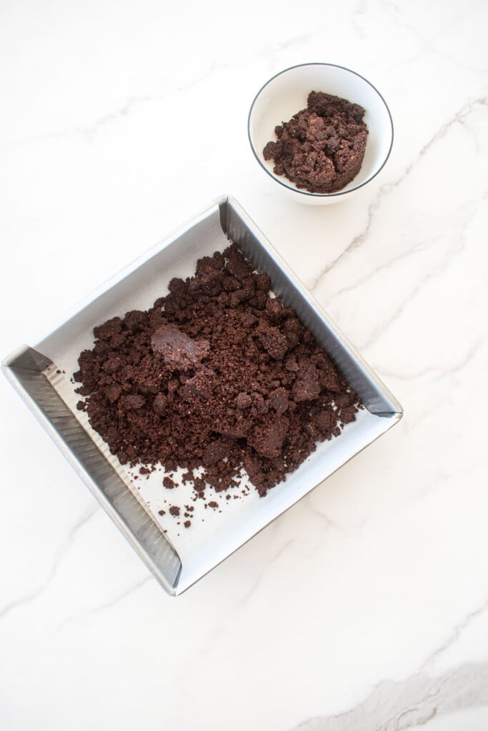 chocolate shortbread dough in an 8-inch baking pan with some set aside in a bowl on a white marble counter.