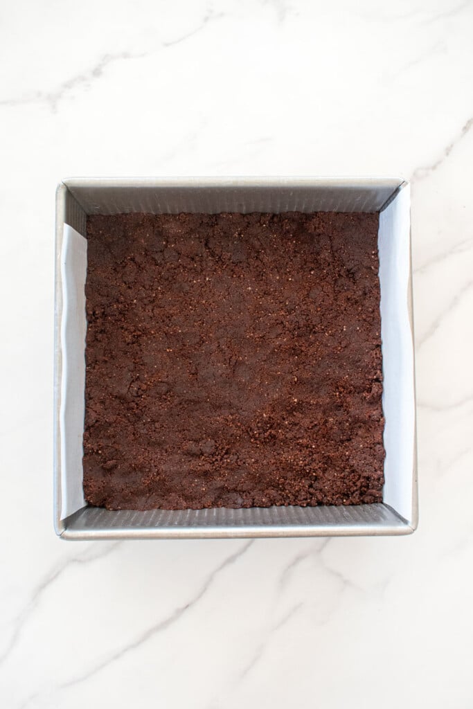 chocolate shortbread dough pressed into a square baking pan on a white marble counter.
