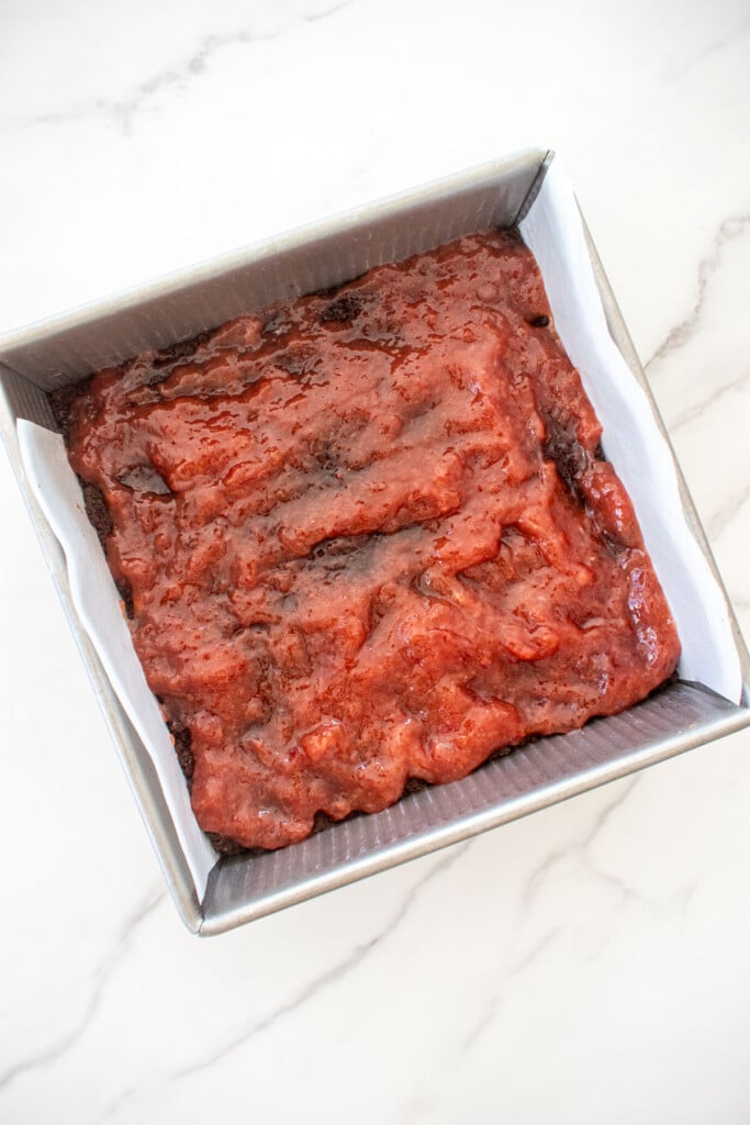 strawberry jam filling on top of a chocolate shortbread crust in a square baking pan on a white marble counter.