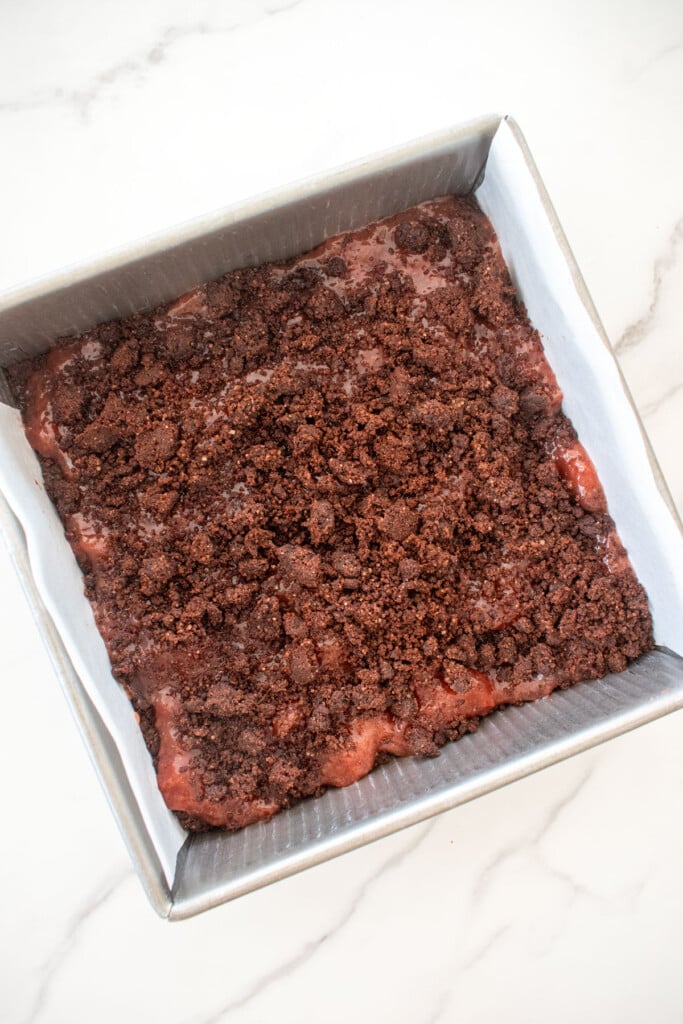 chocolate shortbread cookie dough crumbled on top of strawberry jam in a baking pan on a white marble counter.