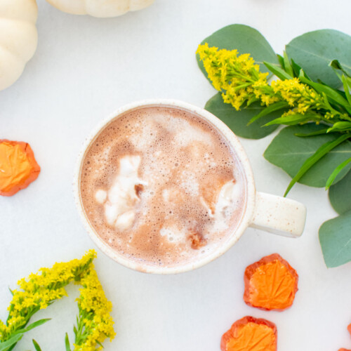coconut hot chocolate with vegan whipped cream in a mug on a white table with fall pumpkins and flowers.