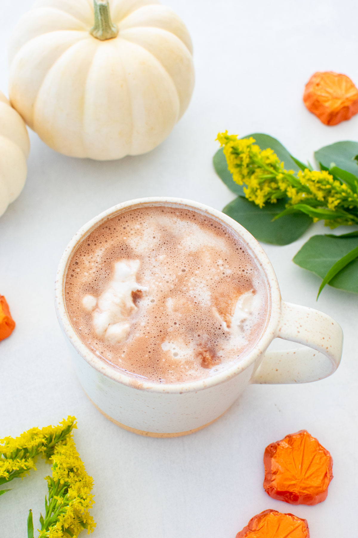 coconut hot chocolate with vegan whipped cream in a mug on a white table with fall pumpkins and flowers.