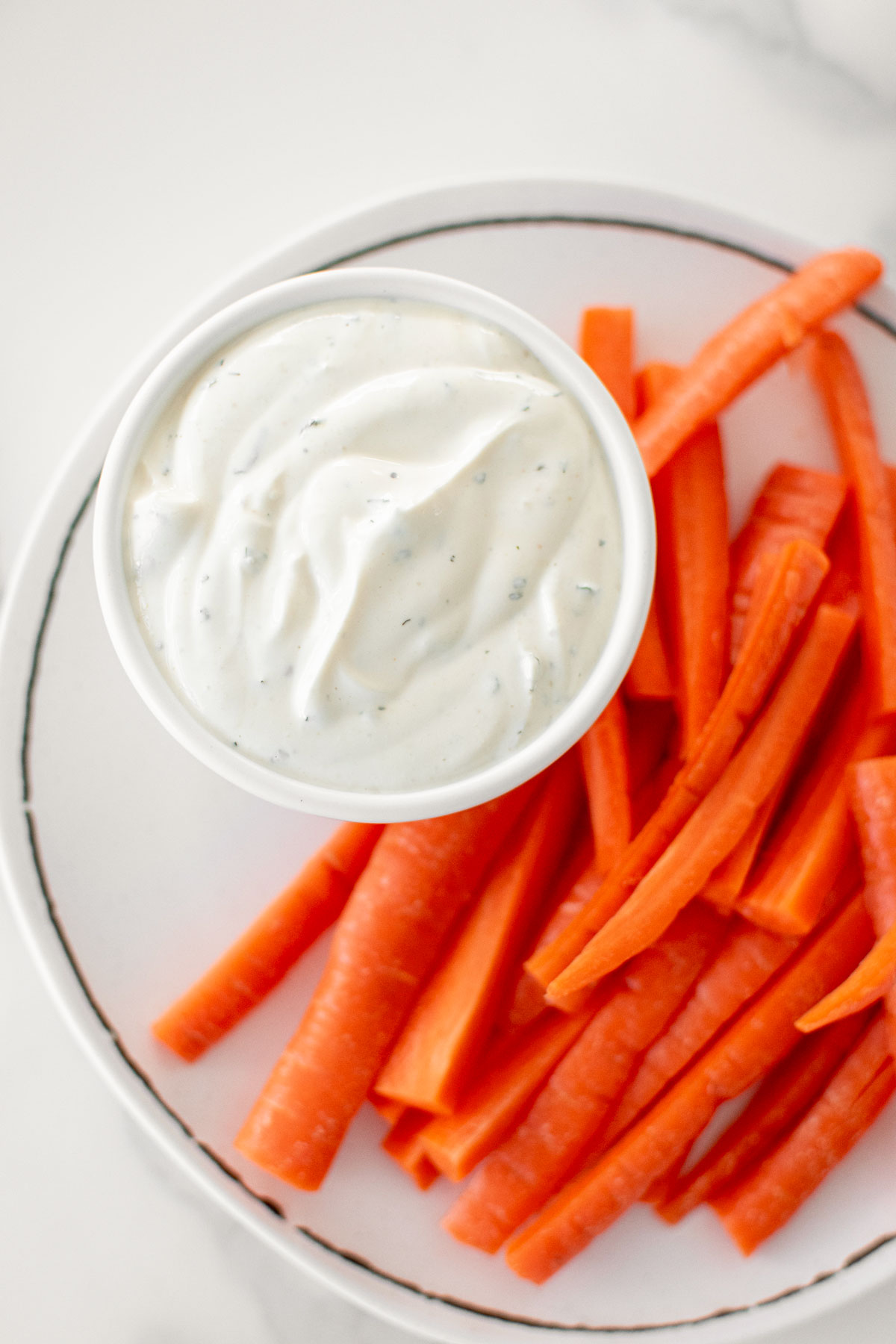 homemade ranch dressing in a dipping dish on a plate with carrots on a white marble countertop.