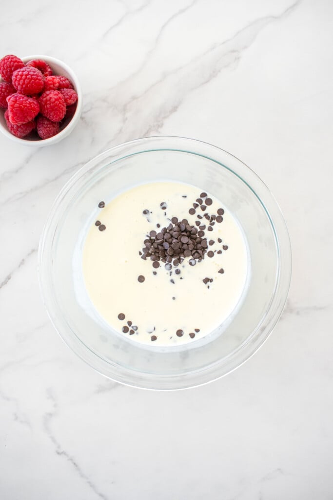 heavy whipping cream and chocolate chips in a glass mixing bowl on a white marble countertop.