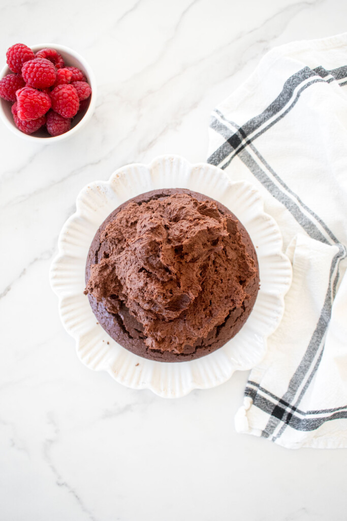 small chocolate cake topped with chocolate ganache frosting on a white plate on a marble countertop.