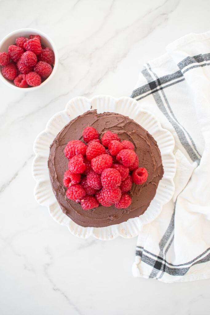small chocolate cake topped with chocolate ganache frosting and fresh raspberries on a white plate on a marble countertop.