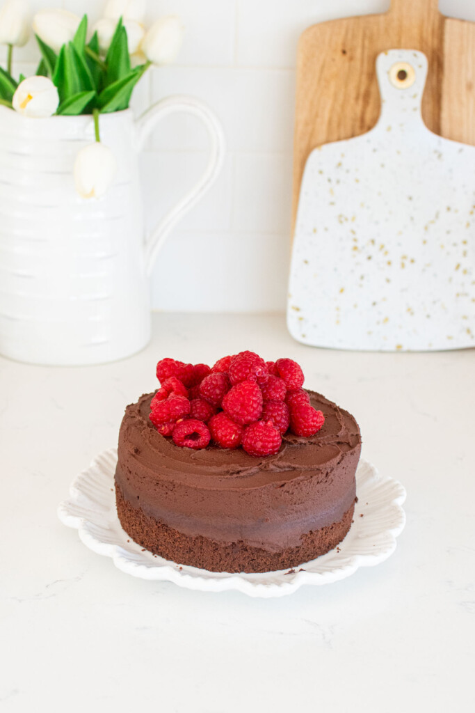 small batch chocolate cake with sourdough discard topped with fresh raspberries on a plate on a white marble kitchen countertop.
