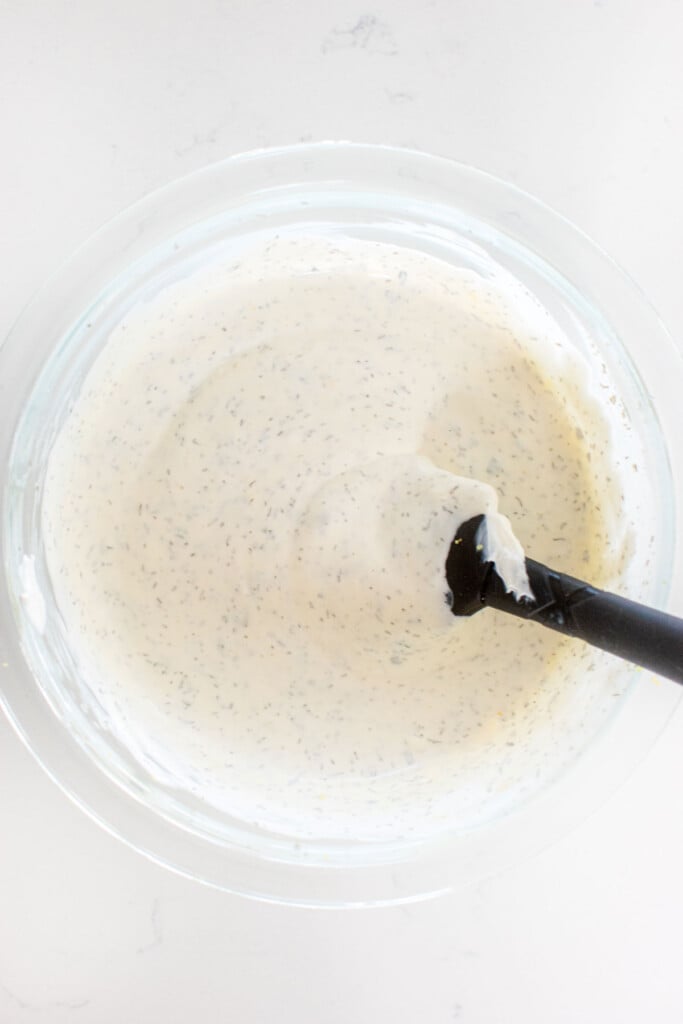 dill dip being mixed together with a spatula in a glass mixing bowl on a marble countertop.