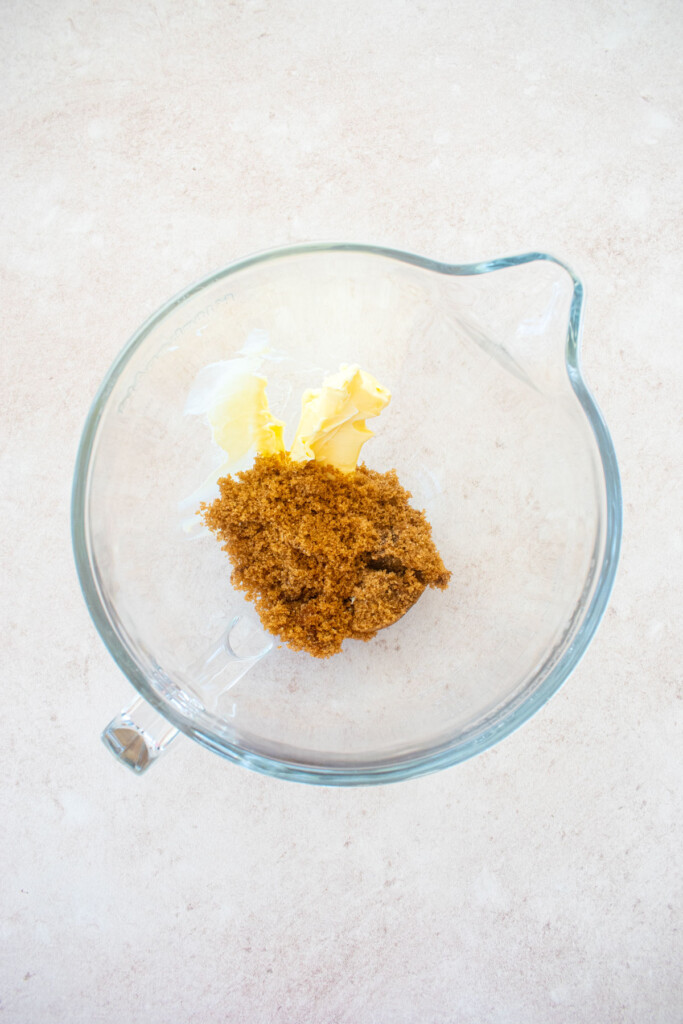glass stand mixing bowl with butter, brown sugar and vanilla on a light pink counter.