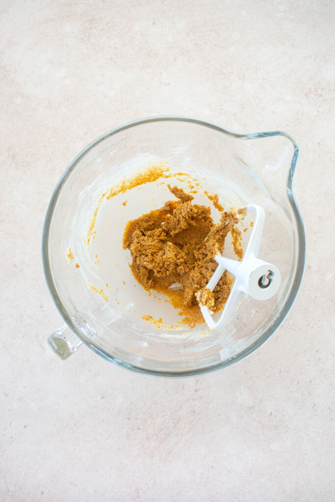 glass stand mixing bowl with creamed butter, brown sugar and vanilla on a light pink counter.