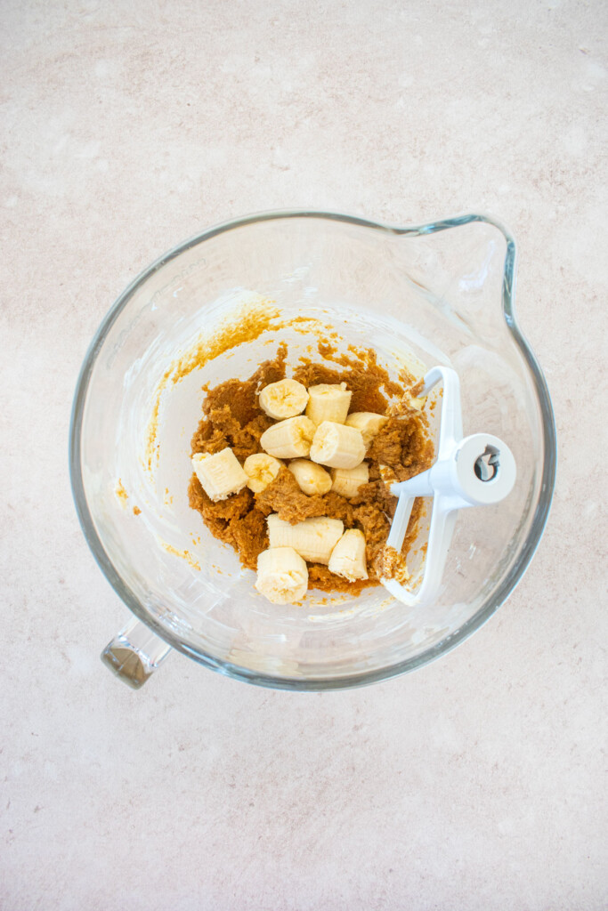 glass stand mixing bowl with creamed butter, brown sugar and vanilla with chunks of banana on a light pink counter.