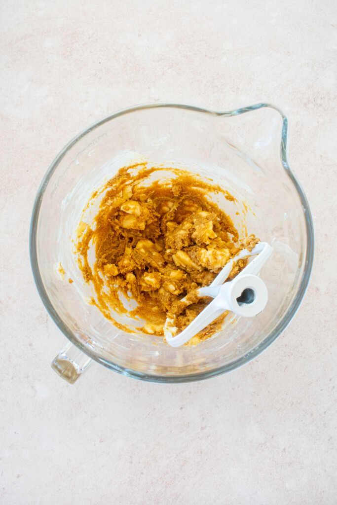 glass stand mixing bowl with creamed butter, brown sugar, vanilla and banana on a light pink counter.