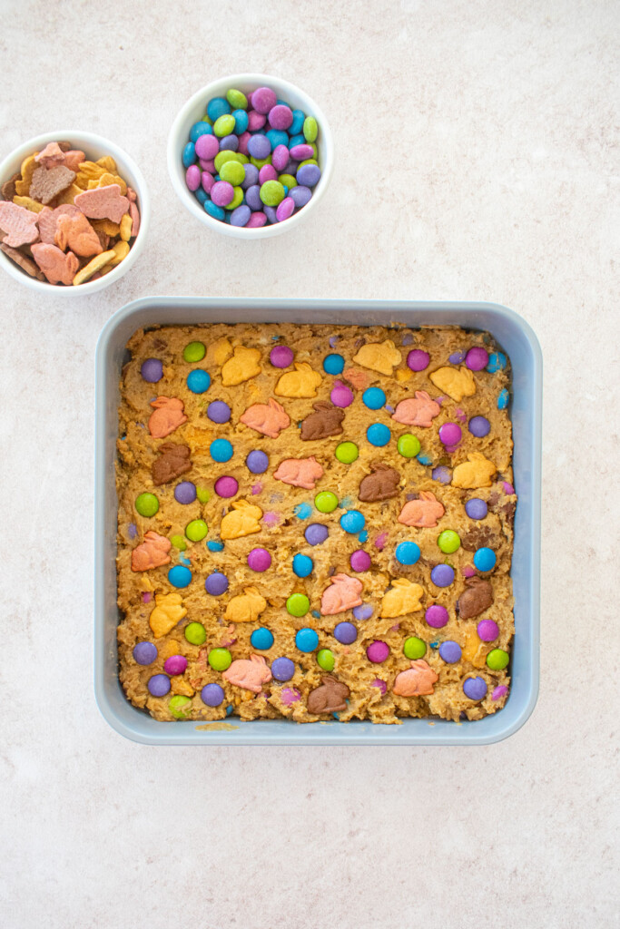 easter banana blondie dough in a square baking pan on a light pink counter with pastel candies and bunnies.