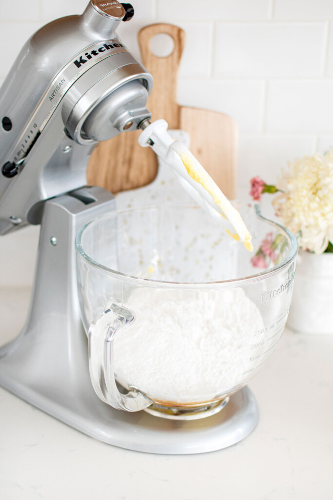 stand mixer with ingredients for honey buttercream frosting in mixing bowl on a white marble counter with fresh flowers.