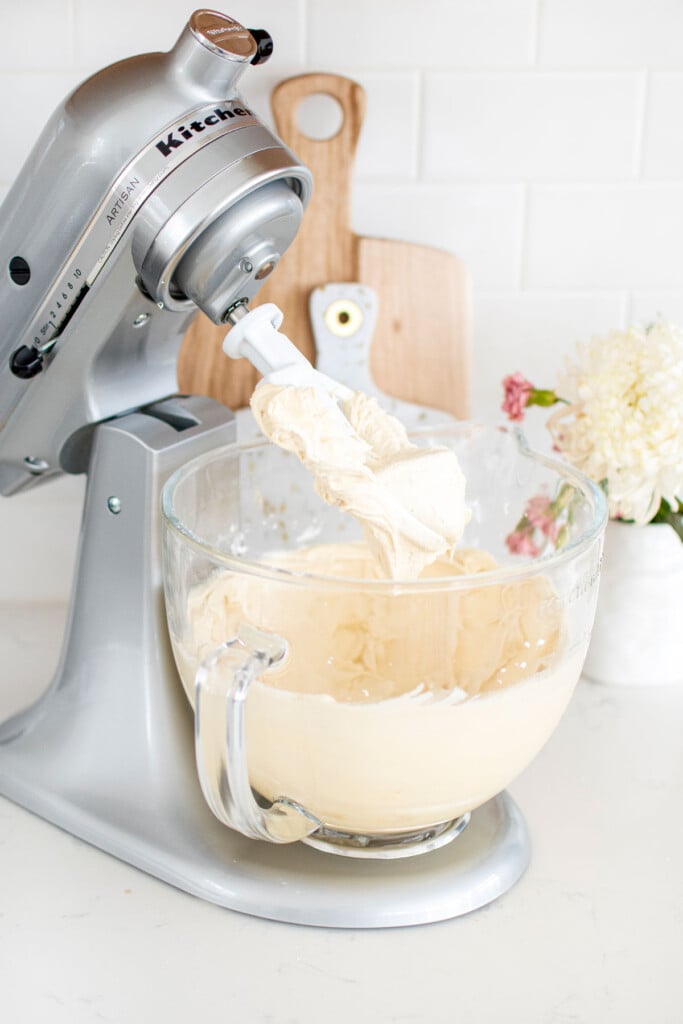 honey buttercream frosting in a glass stand mixing bowl on a white marble counter.