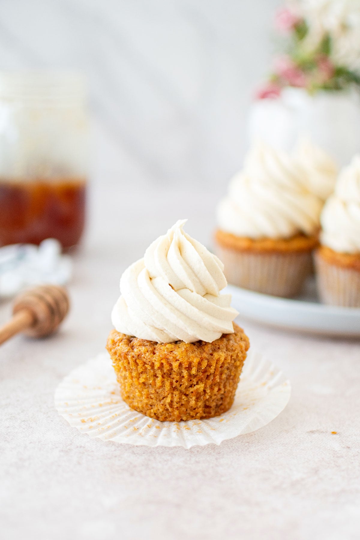 carrot cupcake with the wrapper unpeeled with honey buttercream on top sitting on a table with fresh flowers.